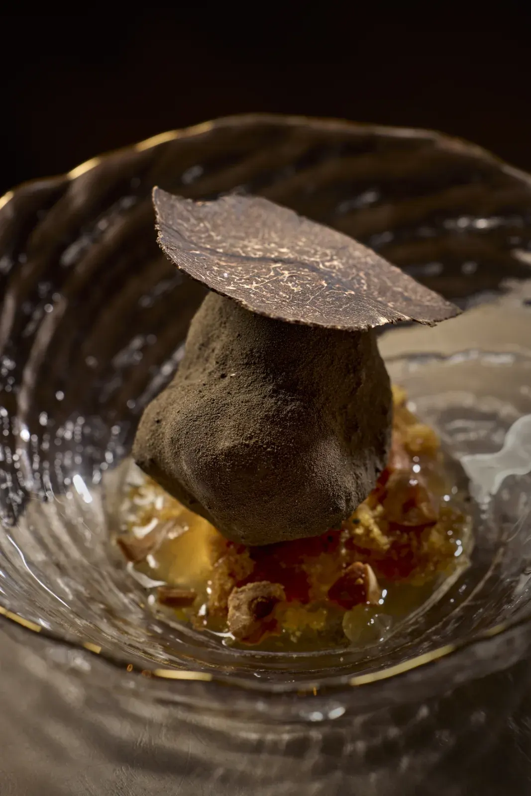 A bite crowned with a slice of truffle, over a golden crumble and its juice, in a glass bowl