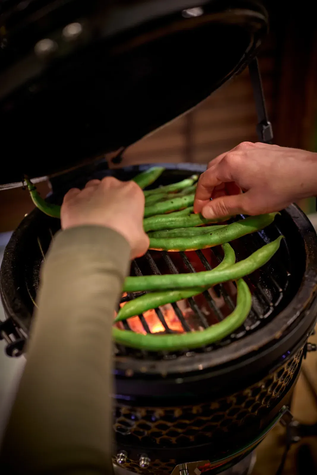 The team's hands placing green pods on the grill, with the embers lit underneath