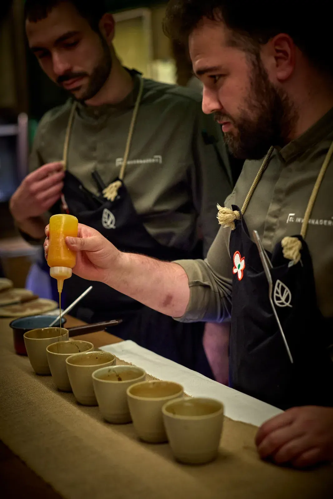 A cook dispenses from a squeeze bottle over a row of ceramic cups lined up on the counter