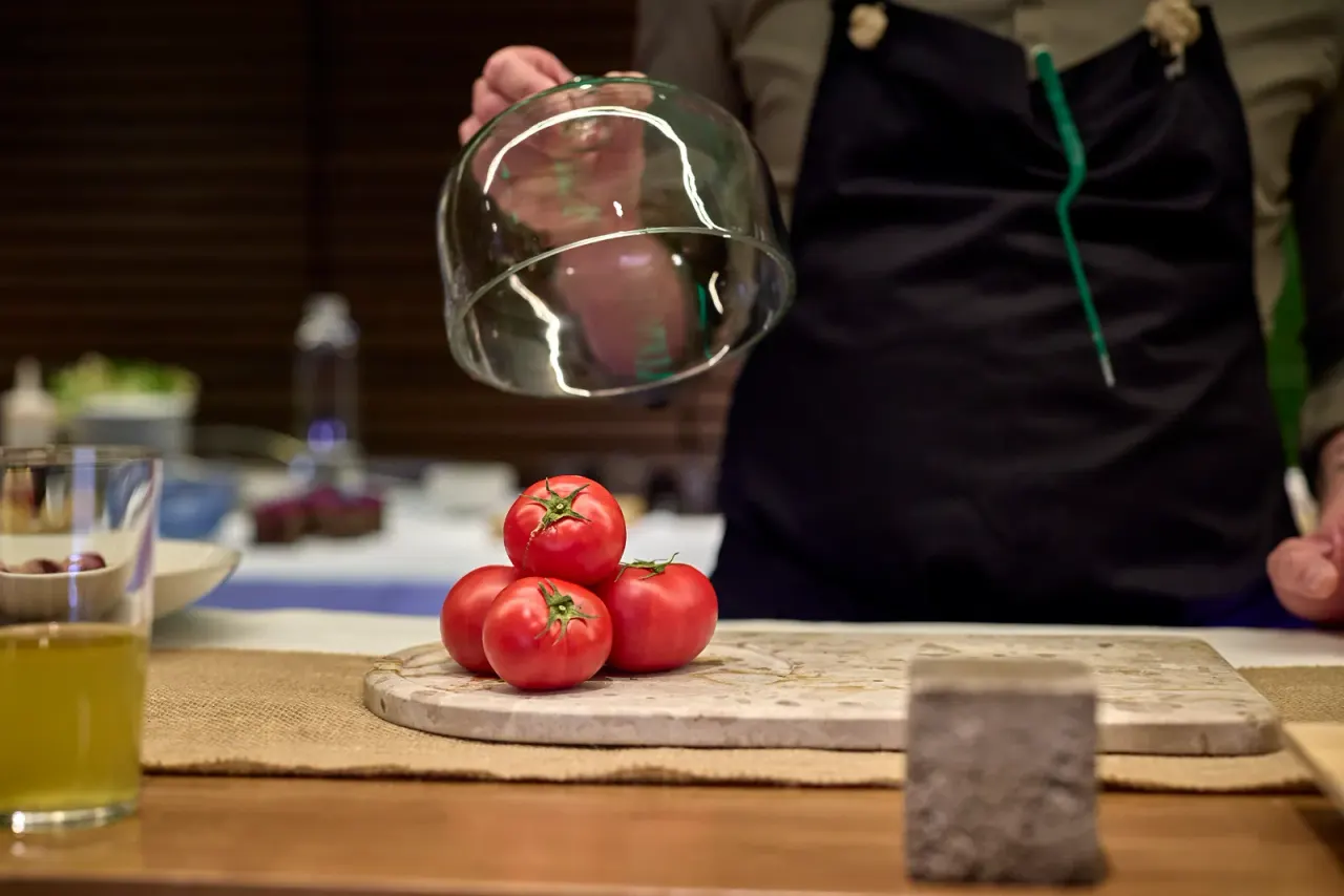Three tomatoes under a glass cloche being lifted by a hand, on a marble disc