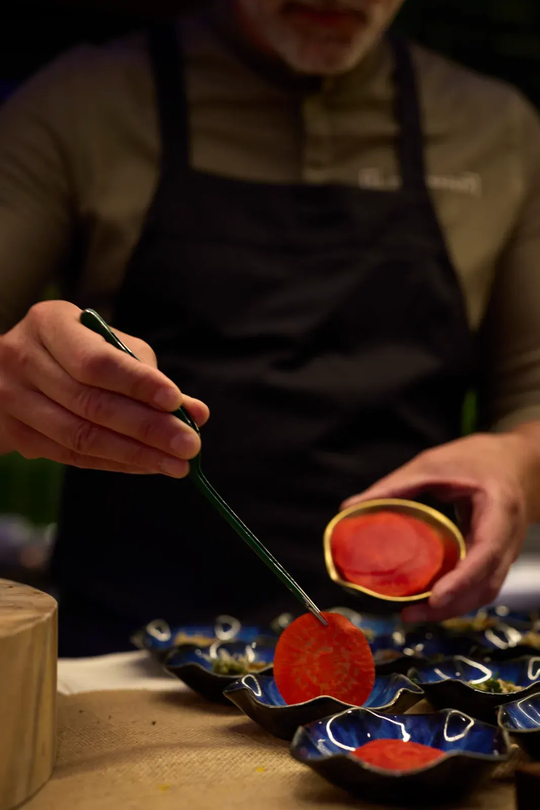 A cook placing red slices with tweezers over a row of blue plates, during plating